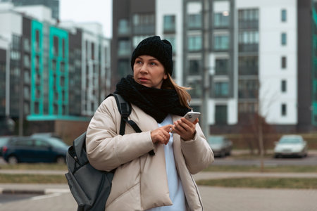 Woman in winter attire checks phone while standing in urban area during cool weatherの写真素材