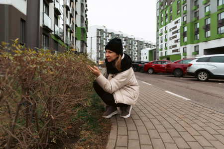 Woman squatting near a hedge in a modern urban neighborhood during an overcast dayの写真素材