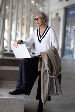 Stylish elderly woman seated indoors, working on laptop while holding a coat in a modern urban setting during daylightの写真素材