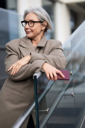 Older woman in stylish glasses leans against glass railing while enjoying a sunny day in an urban environmentの写真素材