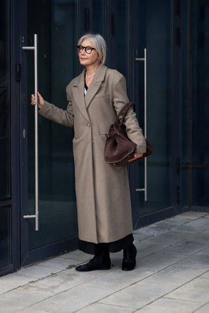 Elderly businesswoman arriving for a meeting in a modern office building, confidently holding a handbag while approaching the entranceの写真素材
