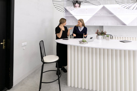 Two women engage in conversation at a modern white reception desk in a sleek office environment during daylightの写真素材