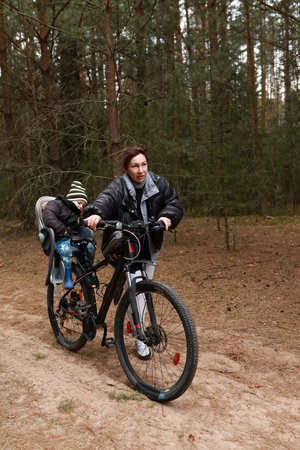 Mother rides bicycle through forest trail with child in seat enjoying outdoor adventure in winter seasonの写真素材