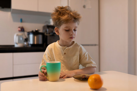 Young boy enjoys morning snack at home while sitting at kitchen table with colorful cup and fruit during bright indoor daylightの写真素材