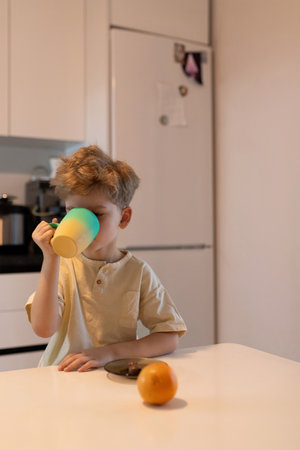 Young boy enjoying a warm drink at a kitchen table in the morning with a fresh orange nearbyの写真素材