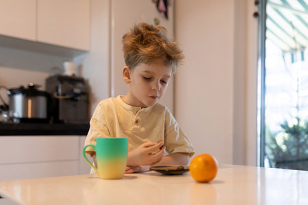 Child enjoying breakfast with orange and beverage while using a tablet in a modern kitchen settingの写真素材
