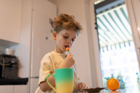 Young boy enjoying a fruit snack while standing at a kitchen counter in a bright home settingの写真素材
