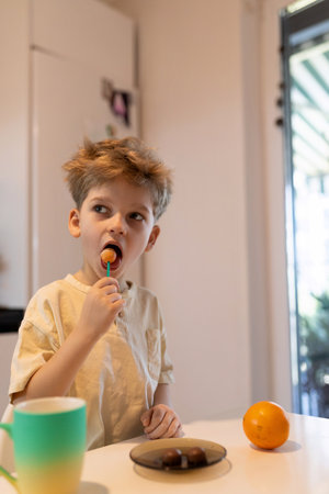 Child enjoys a lollipop while sitting at a kitchen table with an orange in a bright indoor setting during the morningの写真素材