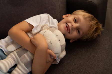 Boy smiles while hugging a soft toy on a cozy couch in a living room during the afternoonの写真素材