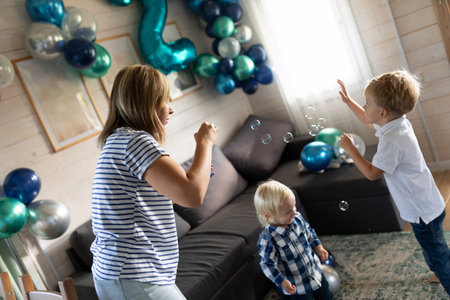 Children celebrate a birthday by playing with bubbles in a decorated living room filled with balloons and festive decorationsの写真素材