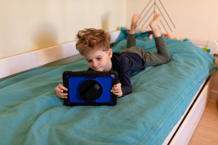 Child engages with tablet on a bed in a cozy room during a sunny afternoonの写真素材