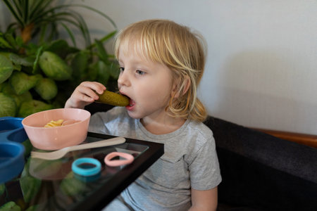 Child enjoys a pickle while sitting at a table with a bowl of pasta in a cozy indoor settingの写真素材