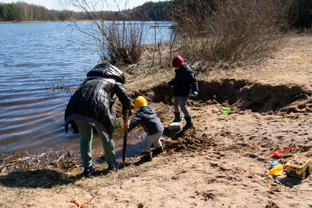 Family playing by the river on a sunny day while children explore the sandy shore and build a small area near the waterの写真素材