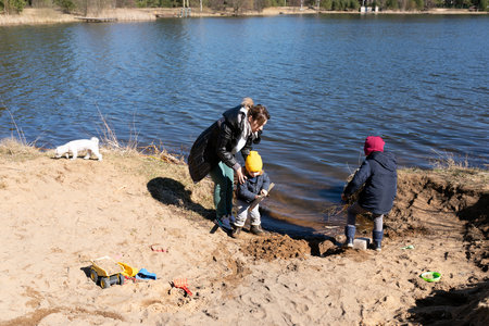 Children play in sand by the lake with an adult supervising on a sunny spring dayの写真素材