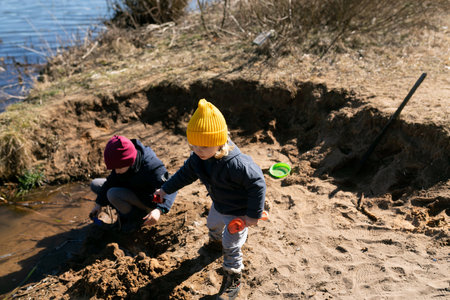 Children playing near the water on a sunny day, digging and exploring the sandy riverbank at a local parkの写真素材