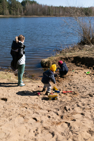 Children play and explore by the lakeside while an adult supervises on a sunny day in early springの写真素材