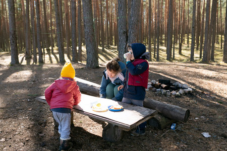 Children enjoy a snack in a serene forest setting with tall trees during a sunny day in autumnの写真素材