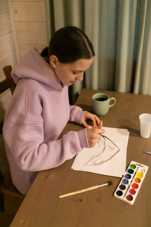 Young artist creates a sketch while enjoying a warm beverage in a cozy indoor setting during the...