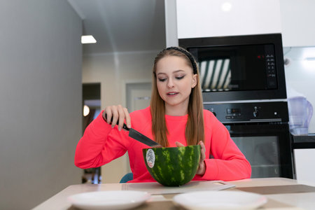 Young girl skillfully prepares watermelon in modern kitchen during daytime activity with friends nearbyの写真素材