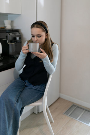 Young woman enjoying a warm drink while seated in a modern kitchen during daylight hoursの写真素材