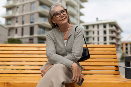 Elderly woman sitting on wooden bench in urban setting, enjoying the outdoors and smiling at the camera during a cloudy afternoonの写真素材