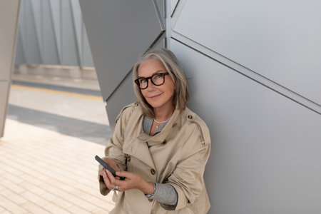 Stylish woman using smartphone while leaning against modern architecture in urban settingの写真素材