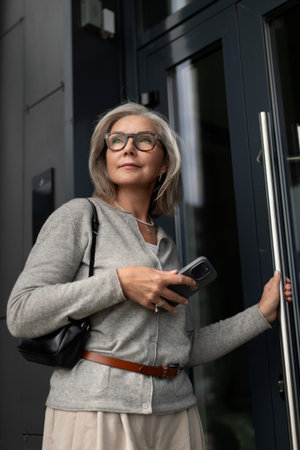 Senior woman entering a modern building while holding a smartphone in an urban setting