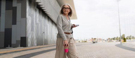 Woman with gray hair holding phone and cup walks along modern architecture in a pleasant urban...