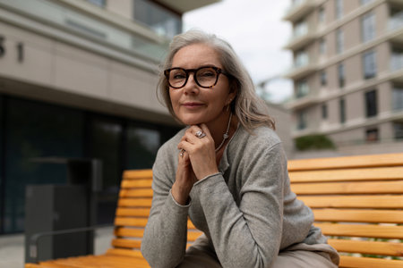 Senior woman enjoys a moment of reflection on a bench in a modern urban setting during a cloudy afternoonの写真素材