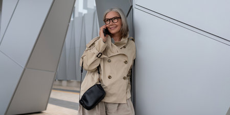 Smiling woman on phone wearing beige trench coat in modern architectural setting during daytimeの写真素材