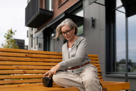 Older woman enjoys coffee on a bench outside a modern building during a sunny dayの写真素材