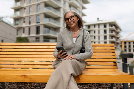 Mature woman smiling while sitting on a bench and using her smartphone in an urban setting on a cloudy dayの写真素材