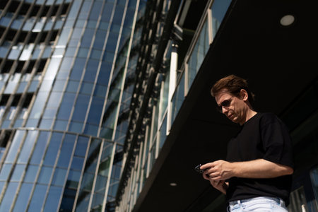 Man using smartphone while standing outside modern building on a sunny day