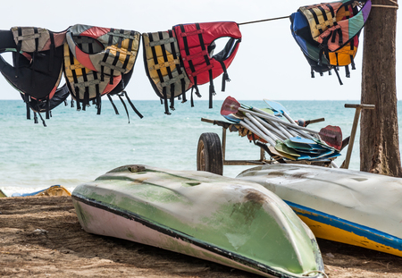 Old life jacket is hanging near the kayak boat and the paddle pile on the beach.の写真素材
