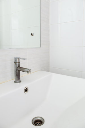 The stained faucet on the white ceramic sink basin near the white tile wall of the restroom in the city house, front view for the background.の写真素材
