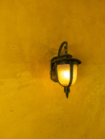 Vintage style metal outdoor lamp glowing on a textured yellow plaster wall background. Traditional Mediterranean architectural lighting.の写真素材