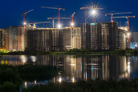 multistory buildings under construction made of reinforced concrete structures and frameworks with cranes at night near pond with reflected lights and glare in water.の写真素材