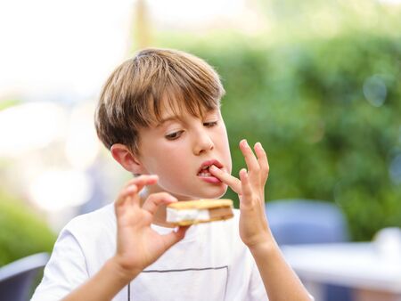 Close up portrait of beautiful boy licking finger while holding and eating sweet ice cream sandwich and looking at ice creamの写真素材