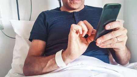 Close view of male patient in recovery after having surgery uses smartphone while lying on a bed in...
