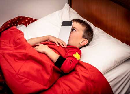 Child under the covers in bed looks at cellphone before falling asleep.の写真素材