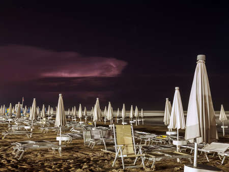 Beach with deck chairs and closed umbrellas on a late summer night. Night thunderstorm seen from the...