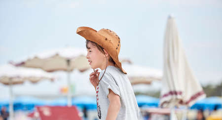 Happy child with cowboy leather hat walking at the beach among the umbrellas. Close-up of a smiling child with cowboy hat, at the beach by the sea.の写真素材