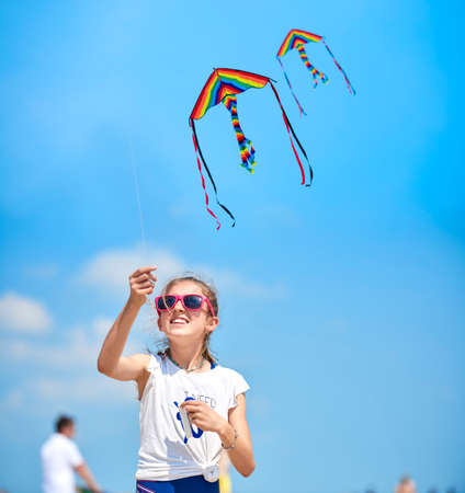 The little girl in the t-shirt plays on the beach by the sea with a colorful kite. In the background many people are walking on the beach.の写真素材