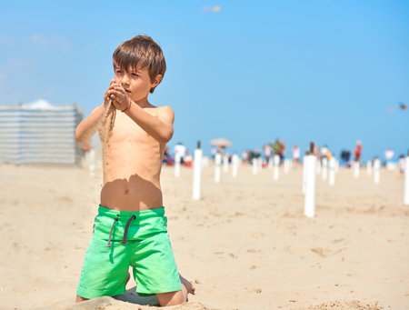 The child in a bathing suit plays with sand at the beach on a sunny day. In the background the beach with people walking by the sea.の写真素材