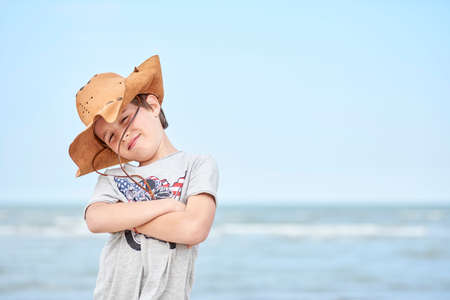 Happy child with cowboy leather hat letting himself be photographed with the sea in the background. Close-up of a smiling child with cowboy hat, at the beach with the ocean in the background.の写真素材