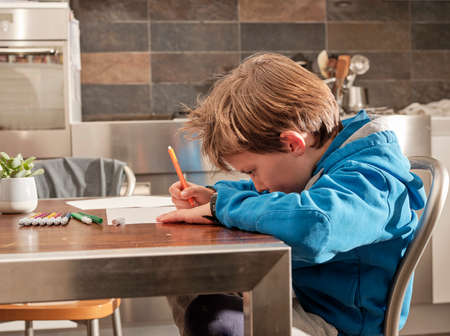 Child draws on a sheet of paper with pencils and markers, sitting at the table in the home kitchen.の写真素材