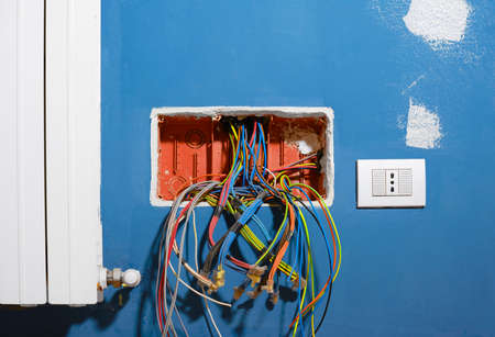 Colored electrical cables coming out of an open junction box, during the renovation of an apartment. Electrical cables coming out of the corrugated pipes inside the junction box located in a blue wall, near a heating radiator and an electrical outlet.の写真素材