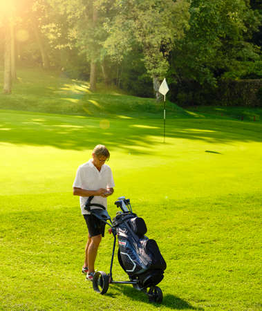 Golfer with cart and bag with golf clubs, holds the score on the golf course at the edge of the green on a sunny day, near the flag.の写真素材