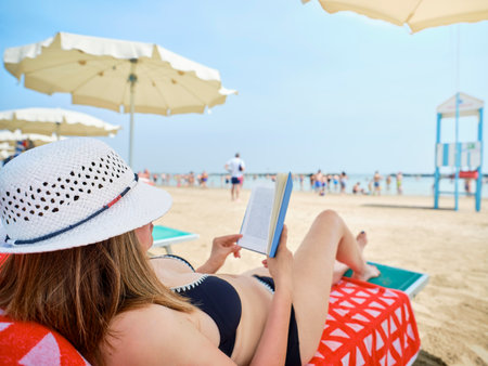Woman wearing a summer hat and lying on a deckchair at the beach while relaxing reading a book, in the background umbrellas, a lifeguard tower and bathers cooling off by the Adriatic sea, Italy.の写真素材
