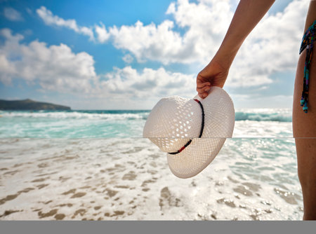Tanned girl holding a white straw hat with her hand, relaxes on the beach looking at the sea on a sunny summer day, in the background the waves of the sea in the Gulf of Policastro, Italy.の写真素材